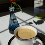 Red rose in blue glass vase beside coffee cup on white table